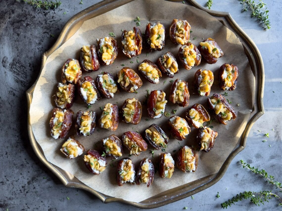A metal tray is covered in parchment paper with rows of goat cheese stuffed dates that have been topped with walnut pieces, thyme, and drizzled in honey.
