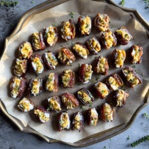 A metal tray is covered in parchment paper with rows of goat cheese stuffed dates that have been topped with walnut pieces, thyme, and drizzled in honey.