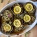 An oval shaped baking dish is full of baked artichokes that have been cut in half and topped with slices of lemon.