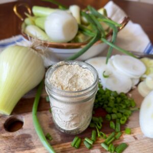 A small jar is full of dehydrated onion powder. Whole fresh onion, slices of onion, and onion greens are surrounding the powder on a wood cutting board.