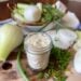 A small jar is full of dehydrated onion powder. Whole fresh onion, slices of onion, and onion greens are surrounding the powder on a wood cutting board.