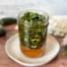 A jar of hot pepper honey sits on top of a small ceramic plate. The jar is filled about 2/3rds full of jalapeno slices and there are a few whole jalapenos garnishing the surrounding area.