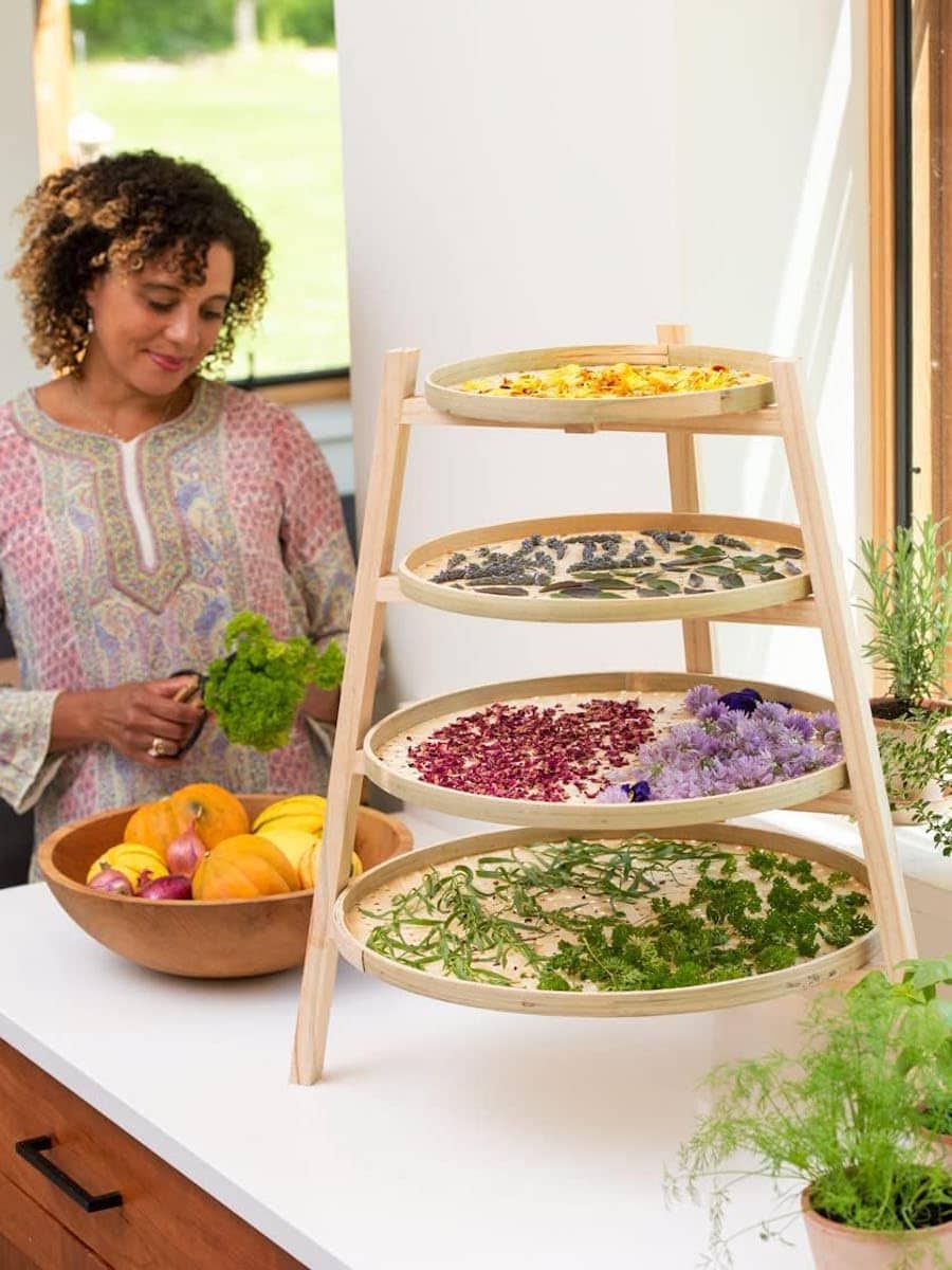 A four tier bamboo herb drying rack on a kitchen counter. The woven trays are round and the bottom is largest, getting smaller as it goes up the four tiers. There are herbs laid out on each of the trays, with a small pot of herbs on the counter nearby. A woman is blurred in the background, sorting through a bowl of herbs