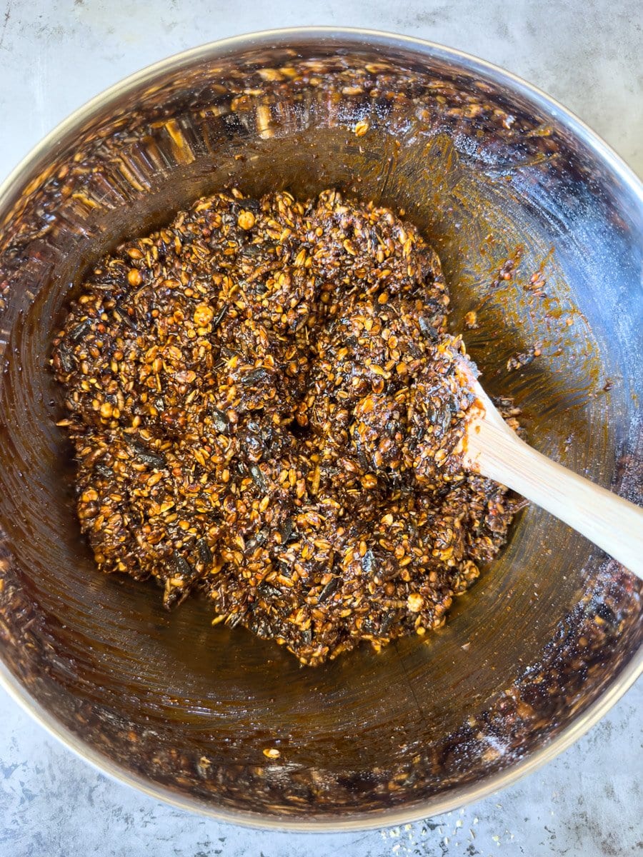 A large metal bowl is full of seeds, chicken feed, grains, and the wet ingredients which creates a wet and semi loose mixture. 