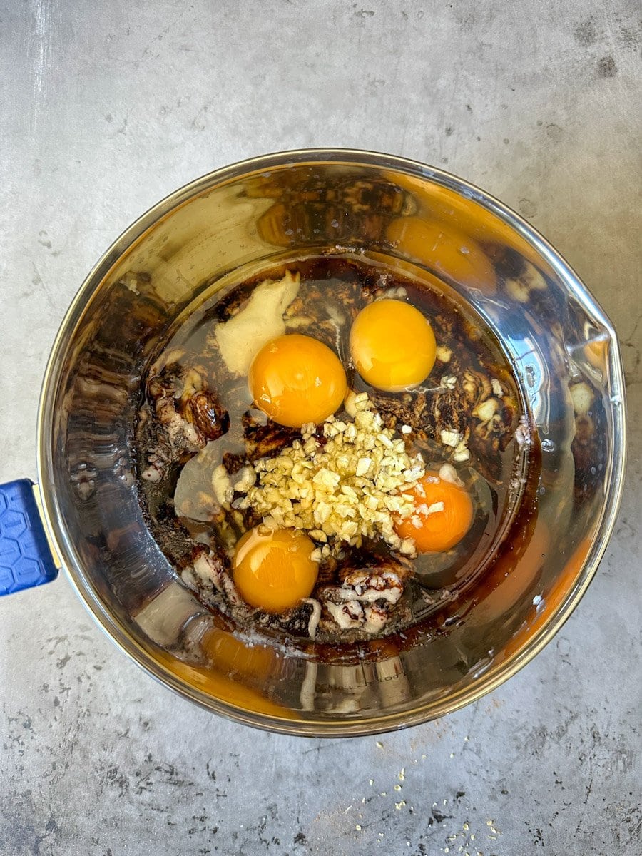 Whole eggs, chopped garlic, coconut oil, and unsulphured black strap molasses make up the wet ingredients inside of a small metal bowl for the flock block recipe. 