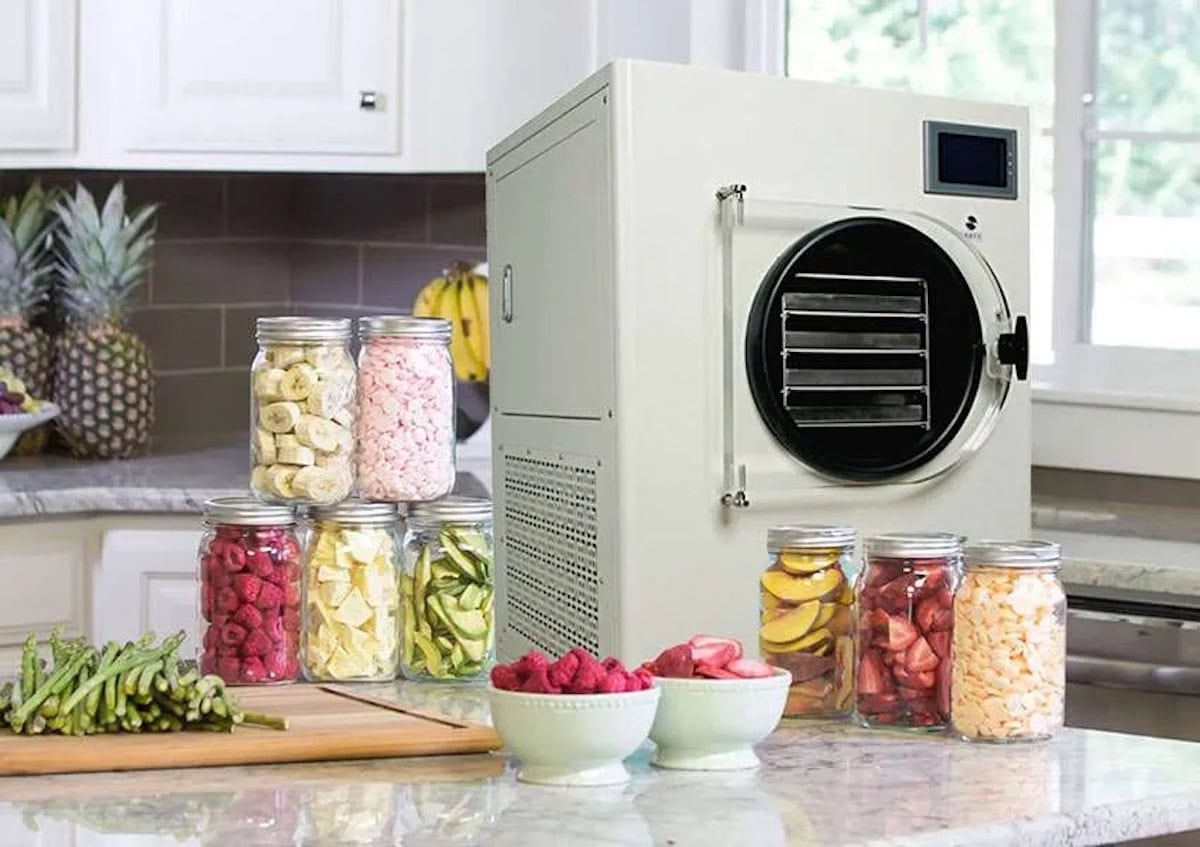 A freeze drier sits atop a countertop with a variety of freeze dried fruits and vegetables packaged into glass quart jars. Some fresh asparagus is on a cutting board while fresh strawberries and raspberries are each in a bowl.