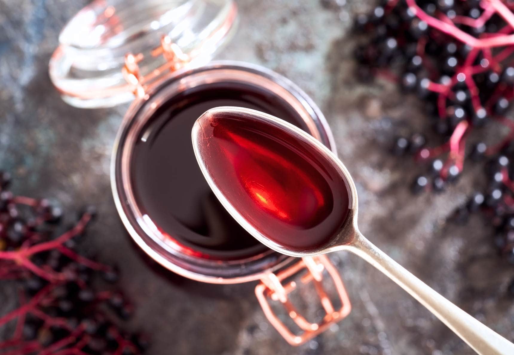 A spoon that is full of elderberry syrup is held over a glass jar with a flip top lid. Fresh berries surround the jar as garnish.