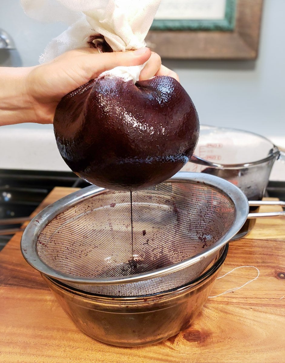 A cheesecloth ball of berries is held above a strainer and glass bowl as its liquid drips into the bowl.