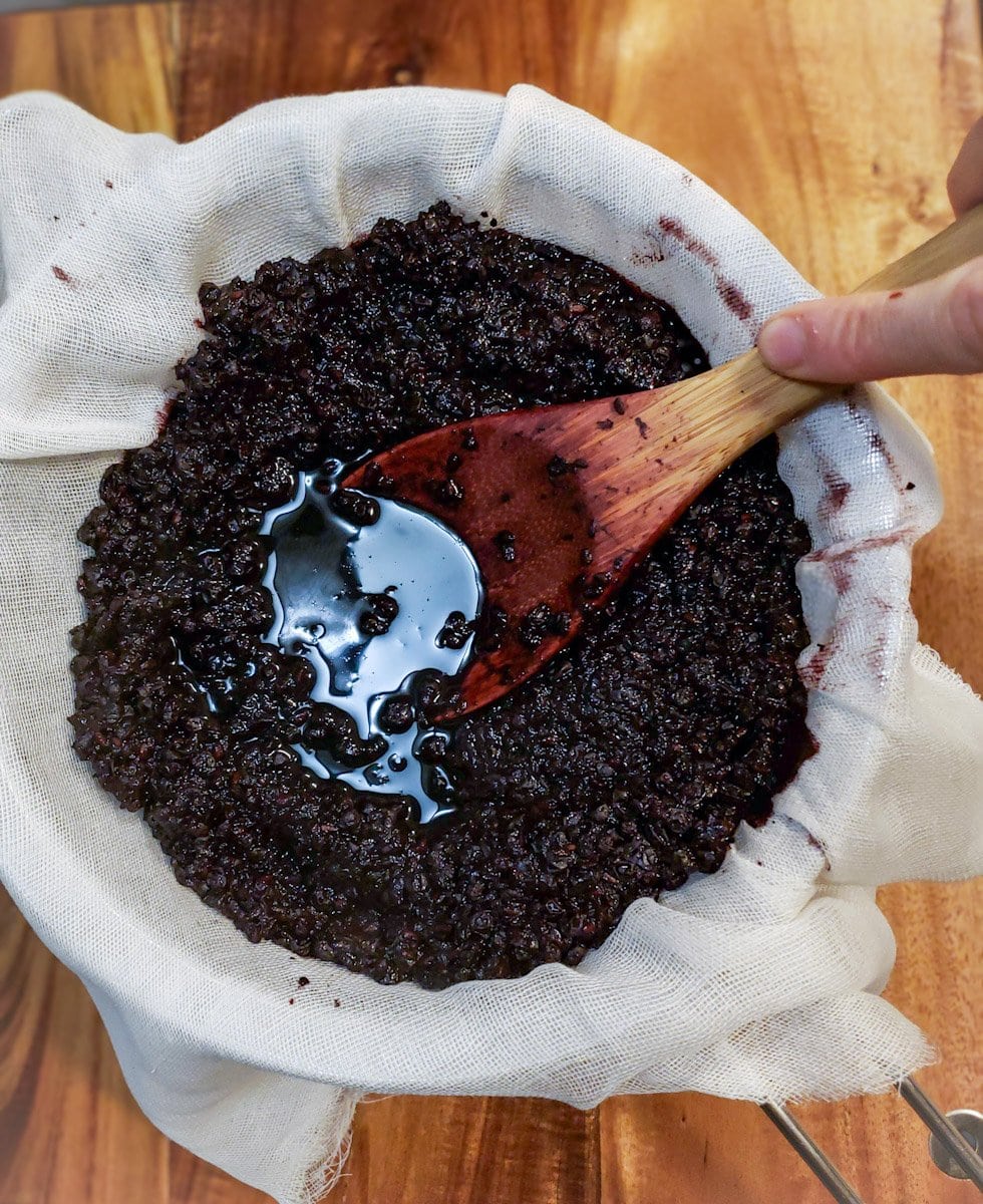 A wooden spoon is being used to push down on the berry and water pulp inside of a strainer that is lined with cheesecloth.
