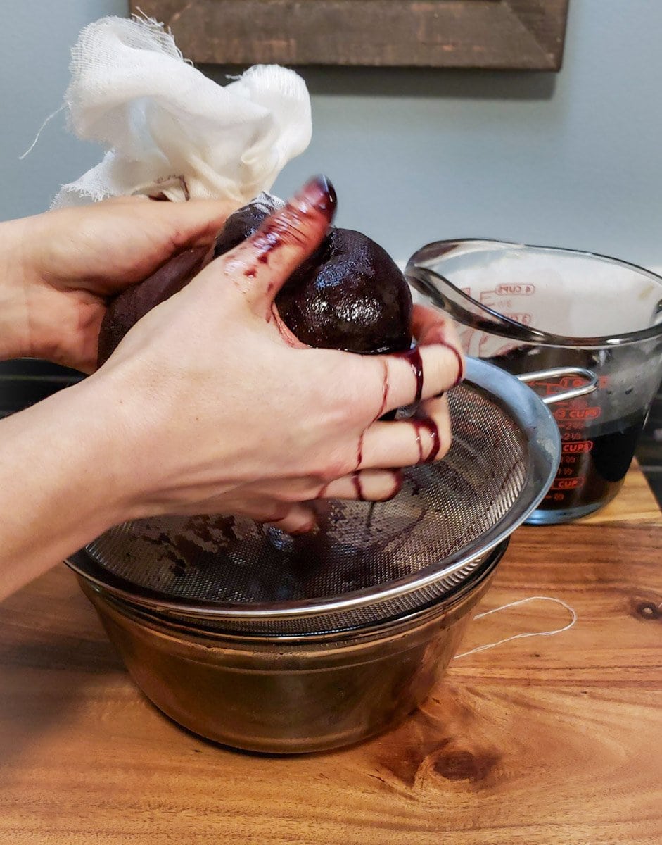 Two hands are squeezing a cheesecloth ball full of cooked berries. Purple juice is running down the hands as it is strained through a mesh strainer into a glass bowl.