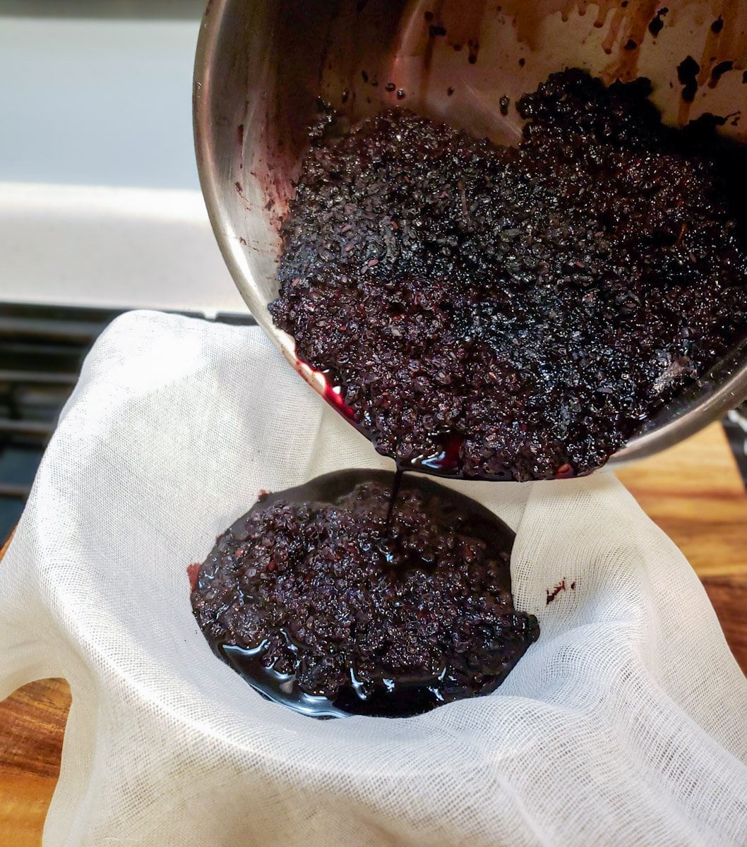 A metal pot of reduced dried berries and water is being poured into a strainer lined with cheesecloth.