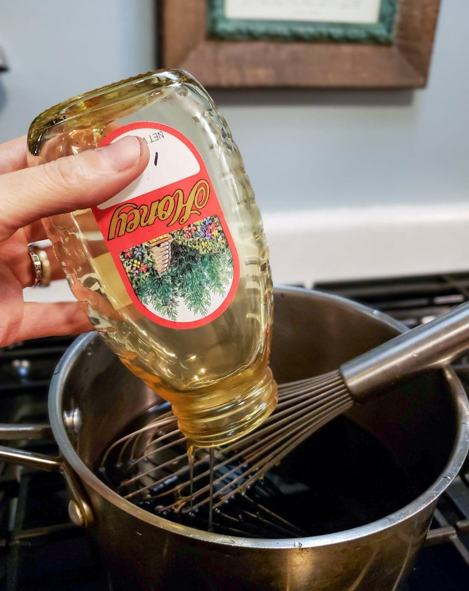 A jar of honey is held upside down to get every last bit of honey into the elderberry syrup inside of a metal pot with a whisk.