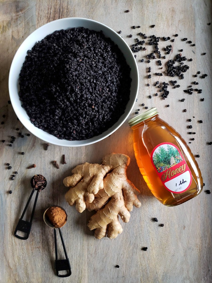 A white ceramic bowl is full of dried berries, loose dried berries, whole cloves, a teaspoon of cinnamon, a hand of ginger, and a bottle of honey surround the bowl.