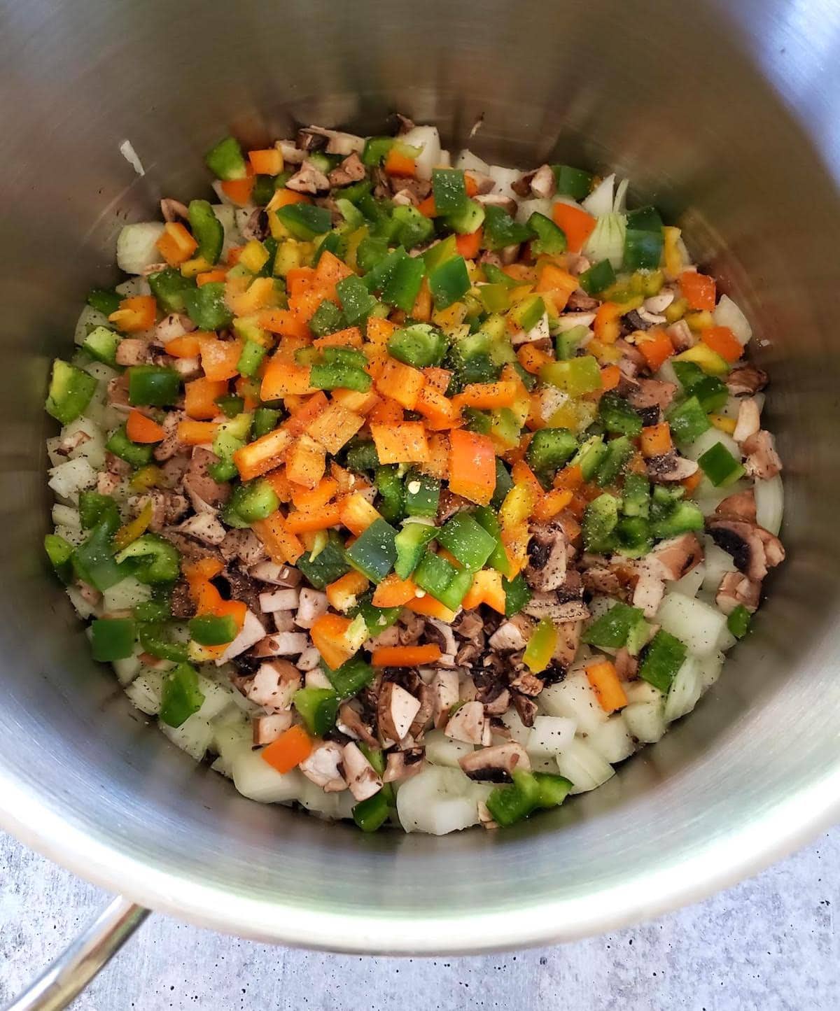 A close up inside a large stainless steel pot, showing chopped onions, mushroom, and bell peppers starting to sauté along with a little salt and pepper.