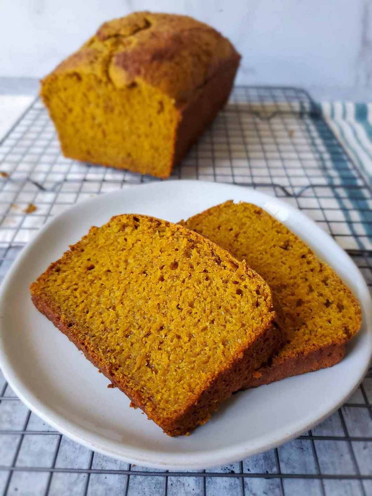 A close up of two slices of sweet bread on a small plate. The bread is slightly orange in color, sourdough pumpkin spice bread. The plate is resting on a cooling rack, and the cut loaf of pumpkin bread is blurry in the background with crumbs scattered around. 