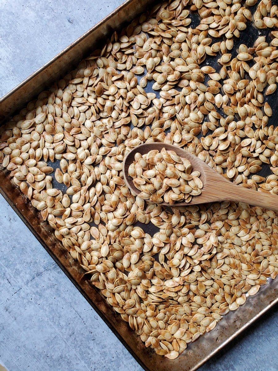 Roasted pumpkin seeds are shown on a baking sheet after they have been roasted. There is a large wooden spoon laying over the baking sheet of seeds with some of the seeds piled onto the spoon. Soaking roasted pumpkin seeds before cooking will make them more nutritious and crunchy. 