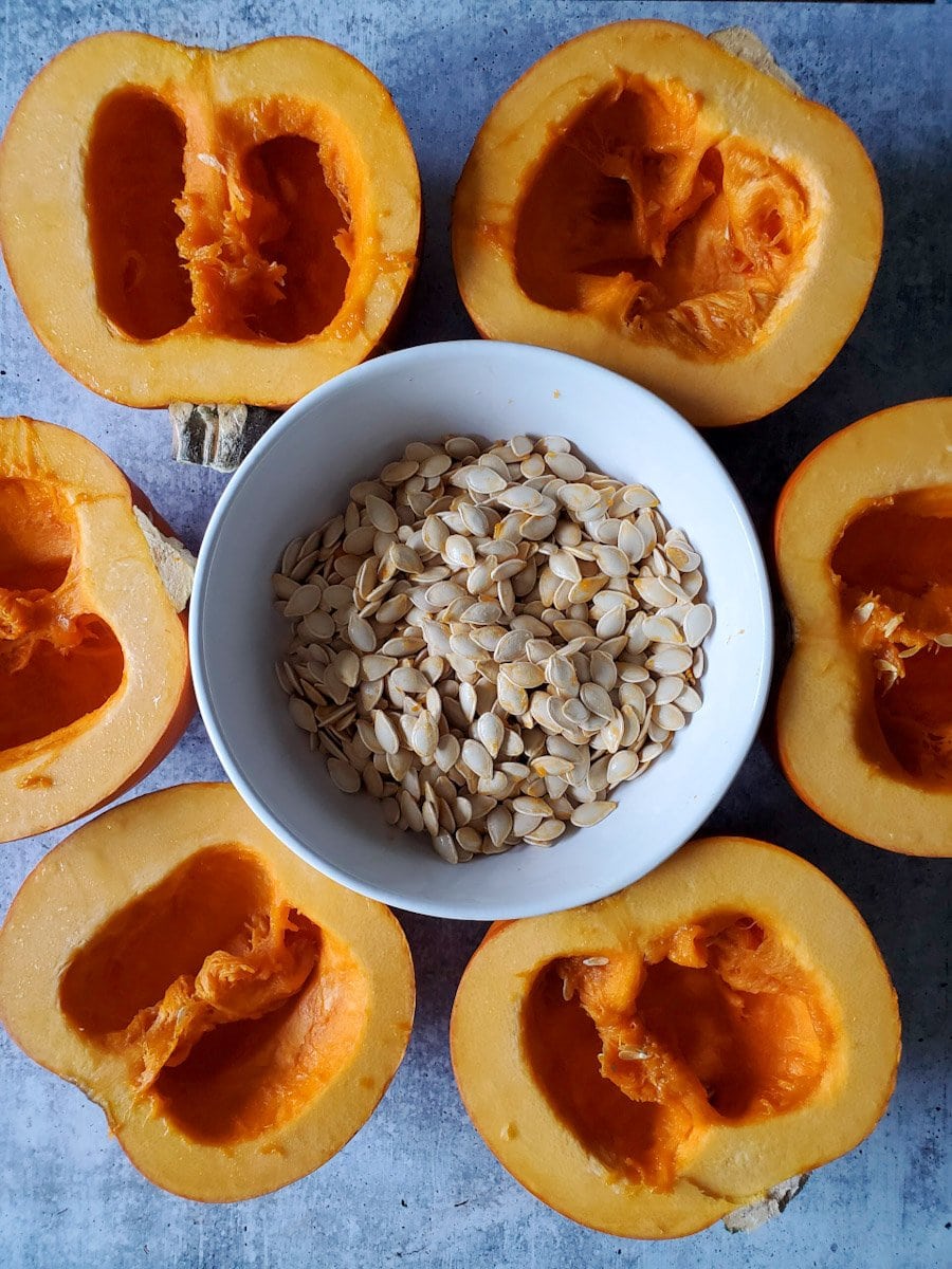 Six halves of sugar pie pumpkins lay flesh side up, their insides are clean of seeds with the remaining guts still left behind. In the middle of the pumpkins lies a white ceramic bowl full of shucked pumpkin seeds. Some of the seeds still have orange pumpkin flesh stuck to them. They will soon be rinsed, strained, and soaked in salt water brine. Soaking roasted pumpkin seeds will help make for a more crunchy and nutritional snack. 