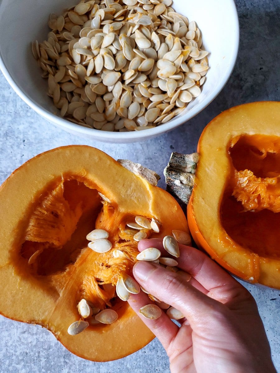 Two sugar pie pumpkin halves are sitting flesh side up and DeannaCat is pulling away some of the seeds from the guts as they are still connected to the inner walls. There is a bowl of shucked pumpkin seeds next to the pumpkins. 