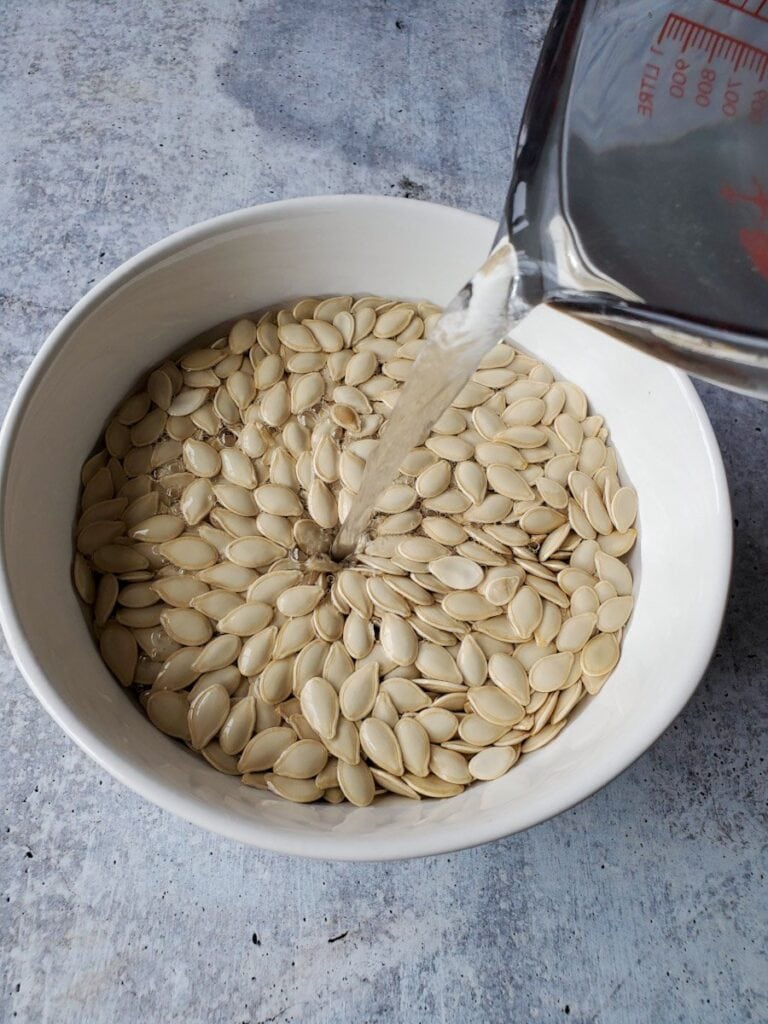 A white ceramic bowl is full of raw pumpkin seeds. A glass measuring cup is pouring a salt water brine into the bowl from the upper right of the image. Soaking roasted pumpkin seeds for 6 to 24 hours is an important step in making the best roasted pumpkin seeds. 