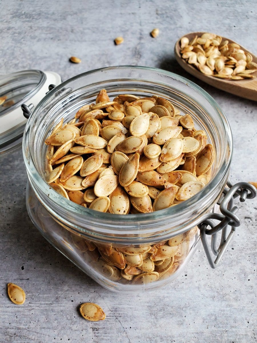 A glass flip top container is shown full to the brim with roasted pumpkin seeds. They are golden brown with specks of salt and paprika. There is a large wooden spoon in the background that has roasted pumpkin flowing over the edges. 