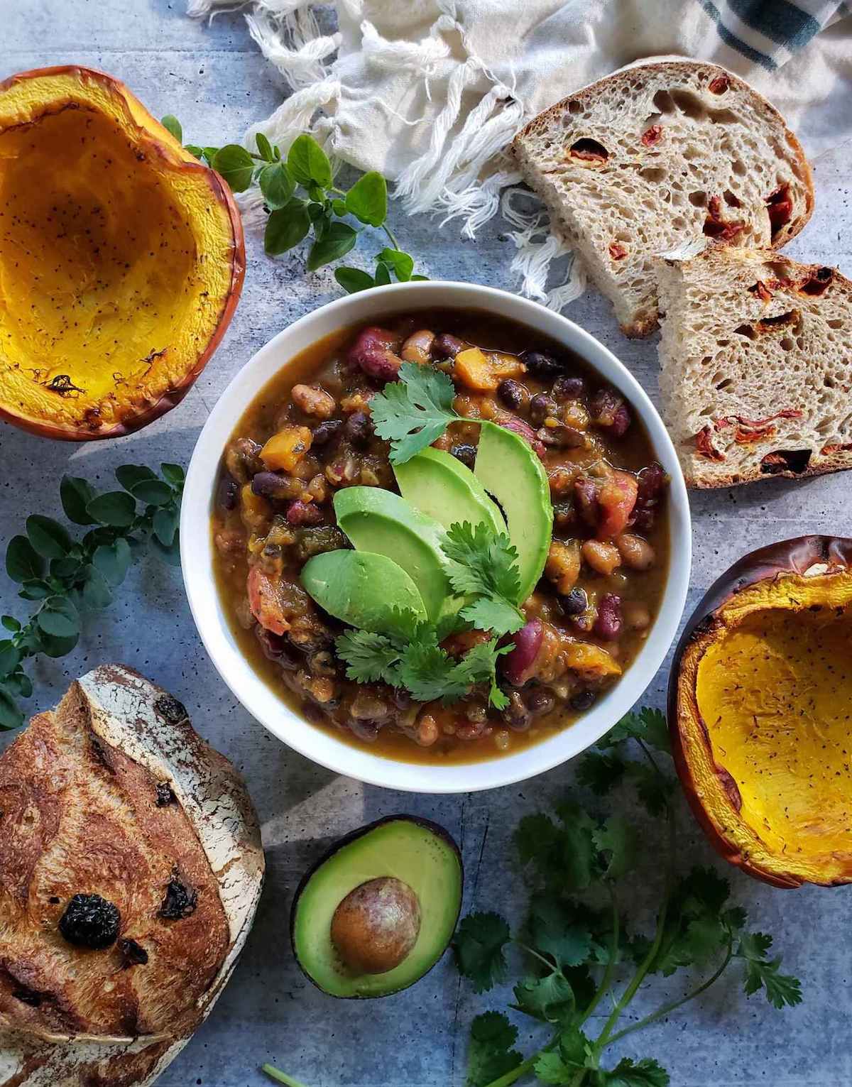 A bowl of vegetarian pumpkin chili topped with avocado slices and cilantro leaves. Surrounding the bowl in a decorative fashion are halves of roasted pumpkin, slices of sourdough bread as well as half of the remaining loaf, half an avocado, sprigs of cilantro, and sprigs of oregano.