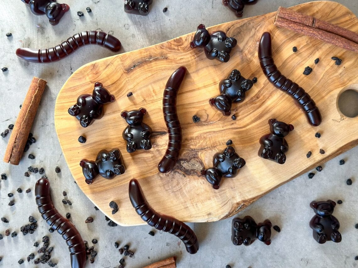 Elderberry gummy bears and gummy worms are arranged on a wooden tray. Gummies, dried elderberries, and cinnamon sticks surround the wooden tray.