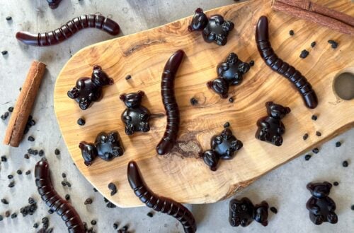 Elderberry gummy bears and gummy worms are arranged on a wooden tray. Gummies, dried elderberries, and cinnamon sticks surround the wooden tray.