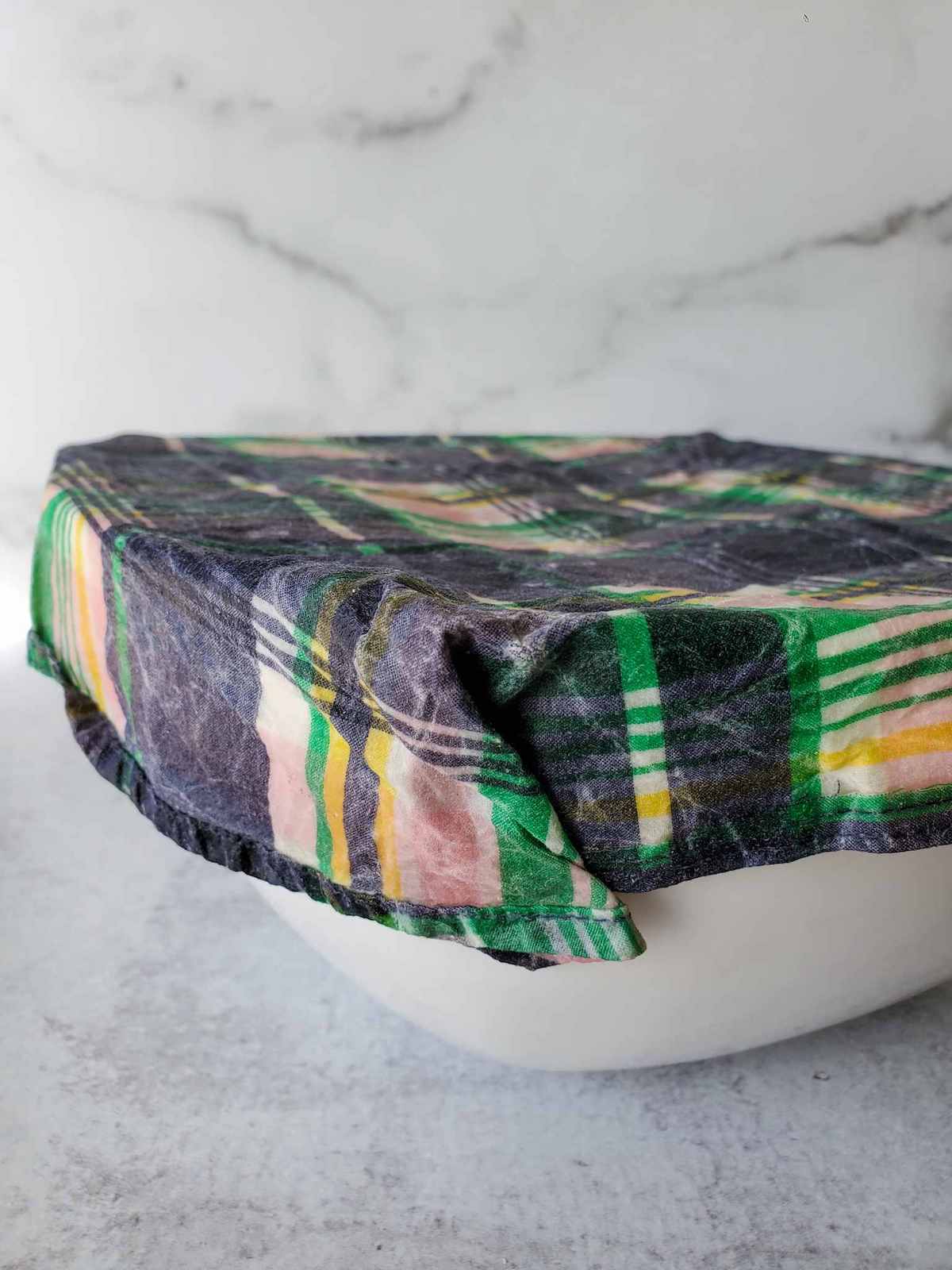 A white ceramic bowl with a plaid color bees wax wrap clinging to the top of a bowl. 