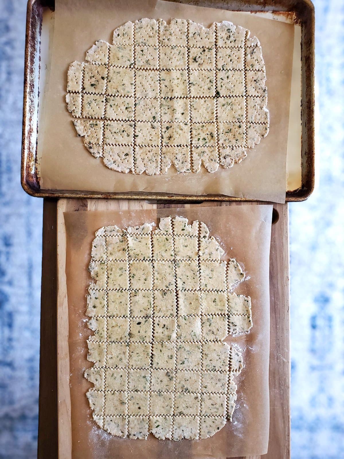 Two baking sheets lined with parchment paper contain dough that has been rolled out thinly and has been cut with a pastry cutter into large, bite sized squares with wavy edges. 