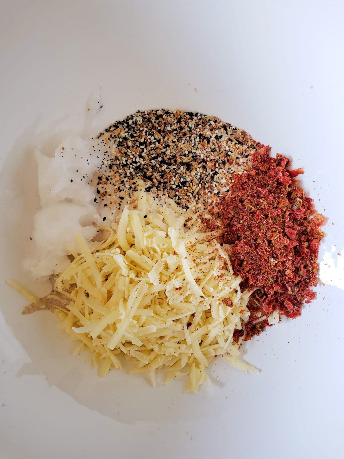 Sun dried tomatoes, seasonings, cheese and coconut oil are inside a large ceramic bowl. 