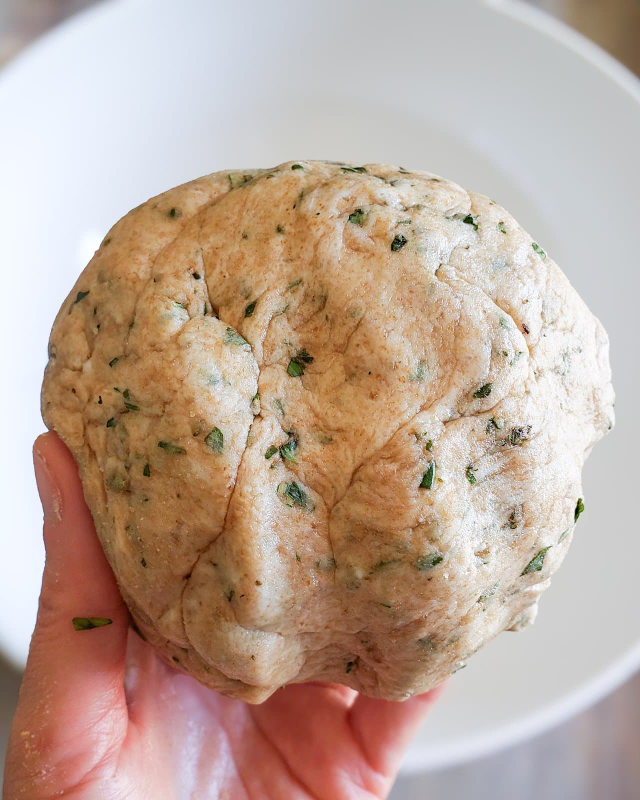 A hand is holding a ball of dough that contains fresh herbs and sun dried tomatoes. 