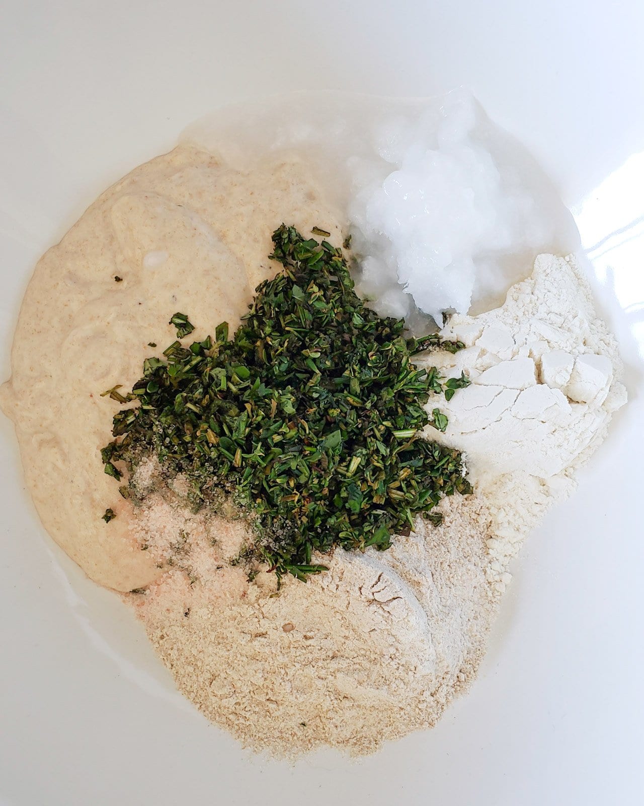 Two types of flour, sourdough starter, coconut oil, and fresh herbs are inside a large white bowl. 