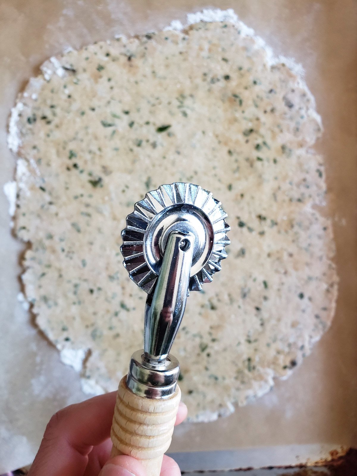 A pastry cutter with a wavy wheel is held above a section of thinly rolled out dough. 