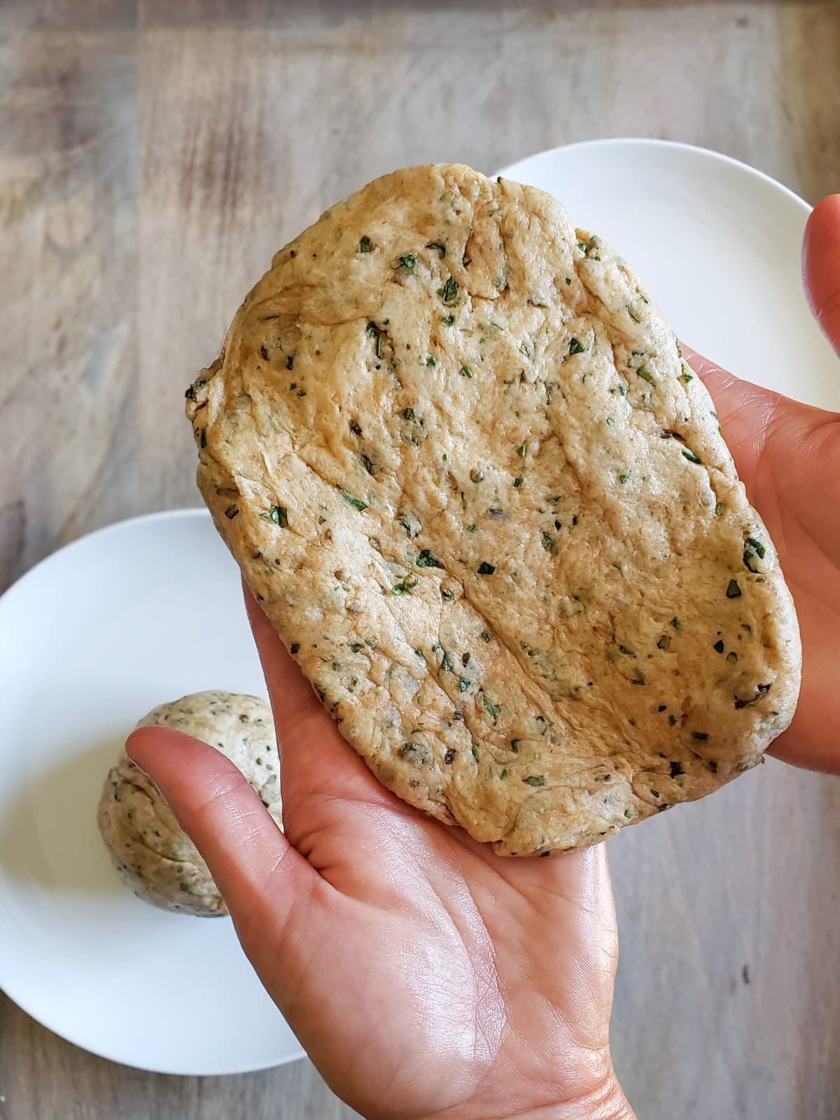 Two hands are holding a freshly shaped sourdough dough ball that is in the shape of a rectangle. Another piece of dough in the shape of a ball sits just below on a plate. 