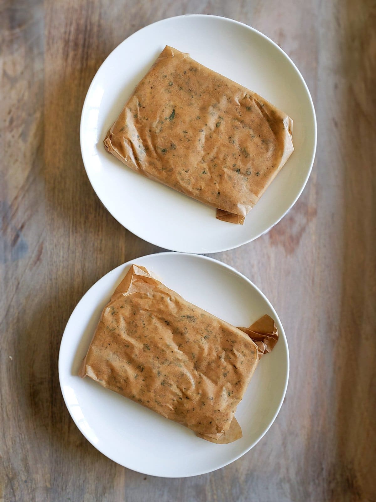 Two ceramic plates contain two rectangles of dough that have been wrapped in parchment paper. 
