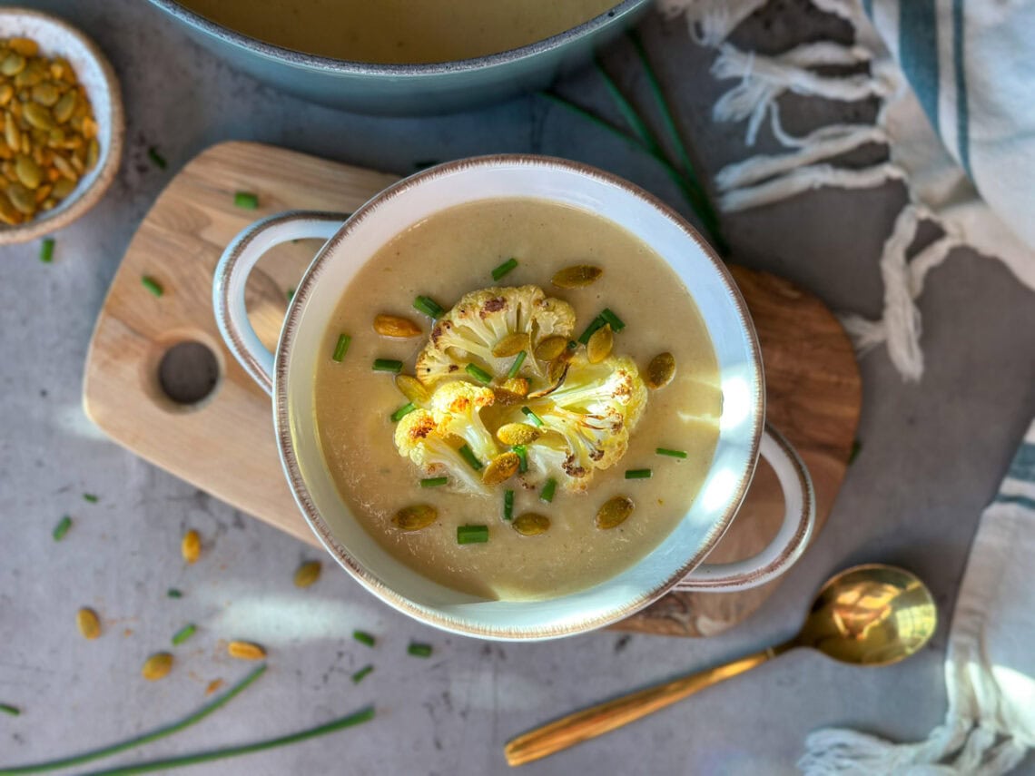 A bowl of roasted cauliflower soup is sitting on a wooden board. It is garnished with a few florets of roasted cauliflower, chopped chives, and seeds. A large blue pot of the soup is sitting just above the bowl.