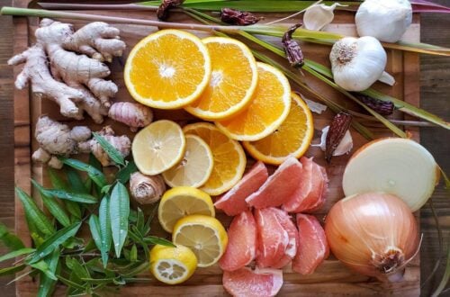 Slices of oranges, lemon, and grapefruit along with whole onion, garlic, ginger, and fresh herbs are on a wood cutting board.