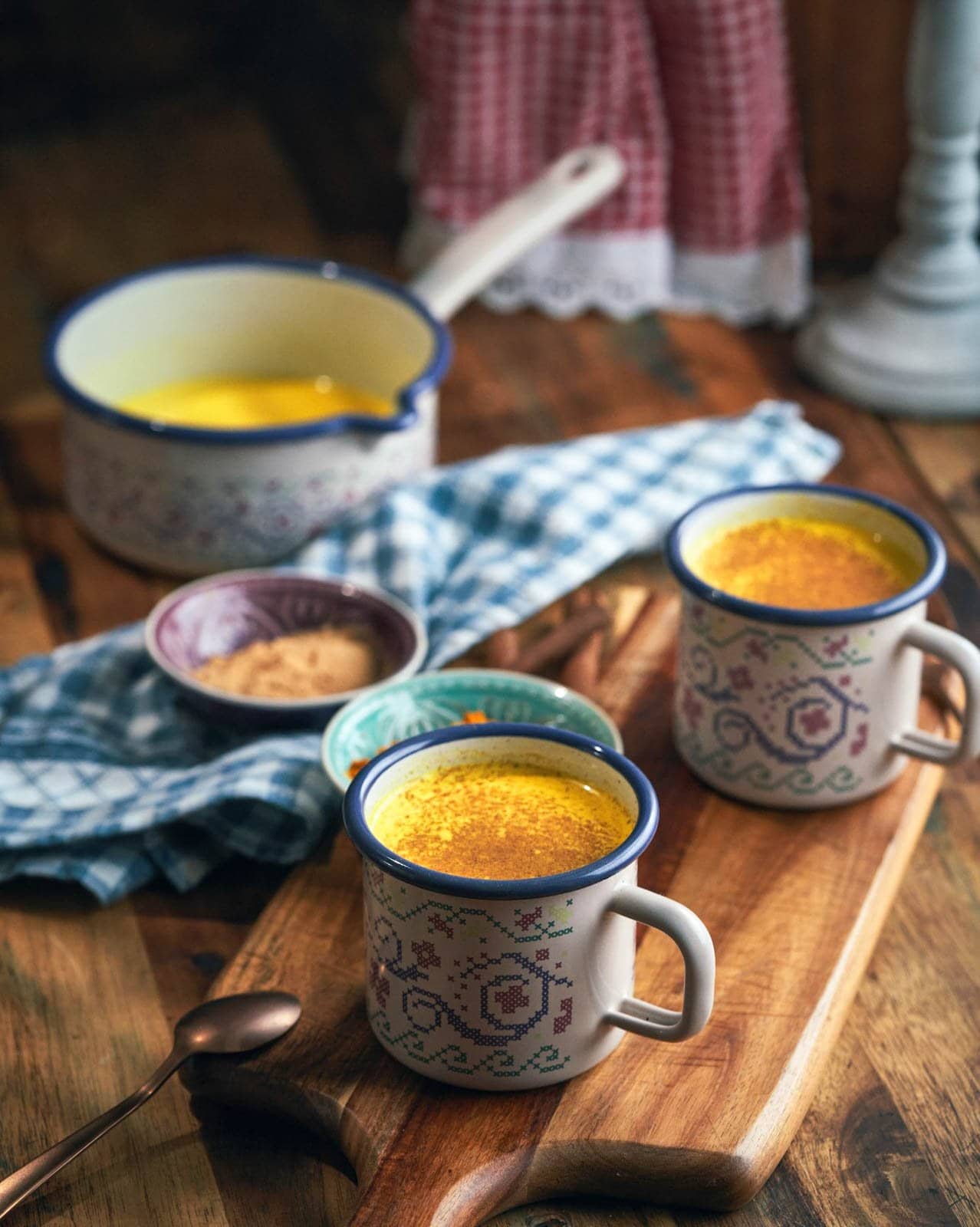 Two ceramic mugs of golden milk garnished with cinnamon powder sit on a wood cutting board. Beyond is a pot of the remaining golden milk.