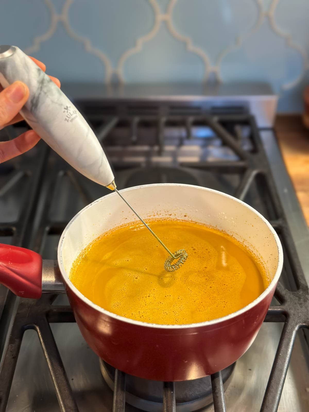 A close up of a small red saucepan on the stovetop, full of bright orange yellow gold milk, with a hand holing an electric whisk frother hovering over the pot