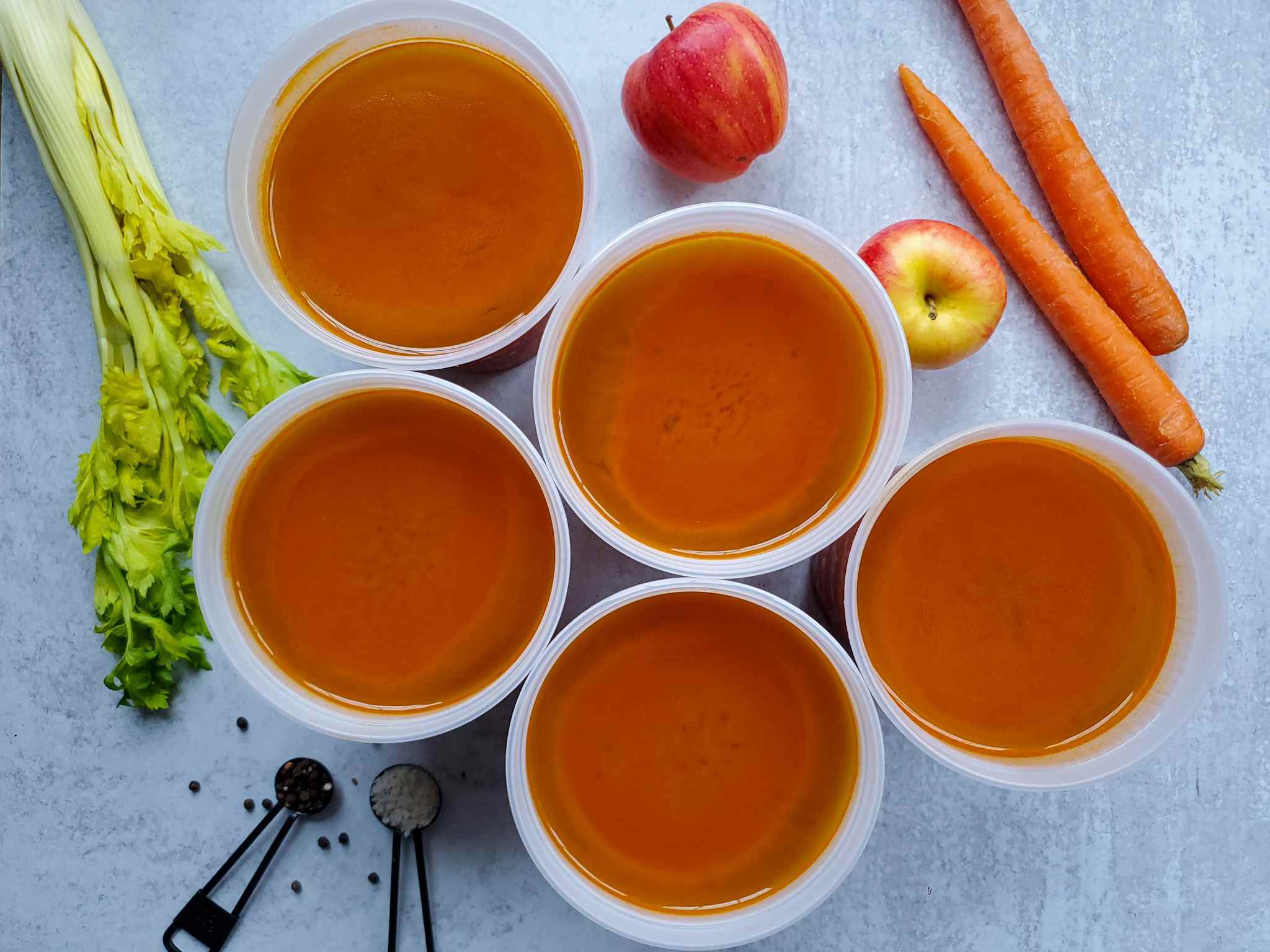 Five quart containers of homemade vegetable broth are arranged in a honeycomb pattern. Celery, carrots, and apple are laid out surrounding the containers.