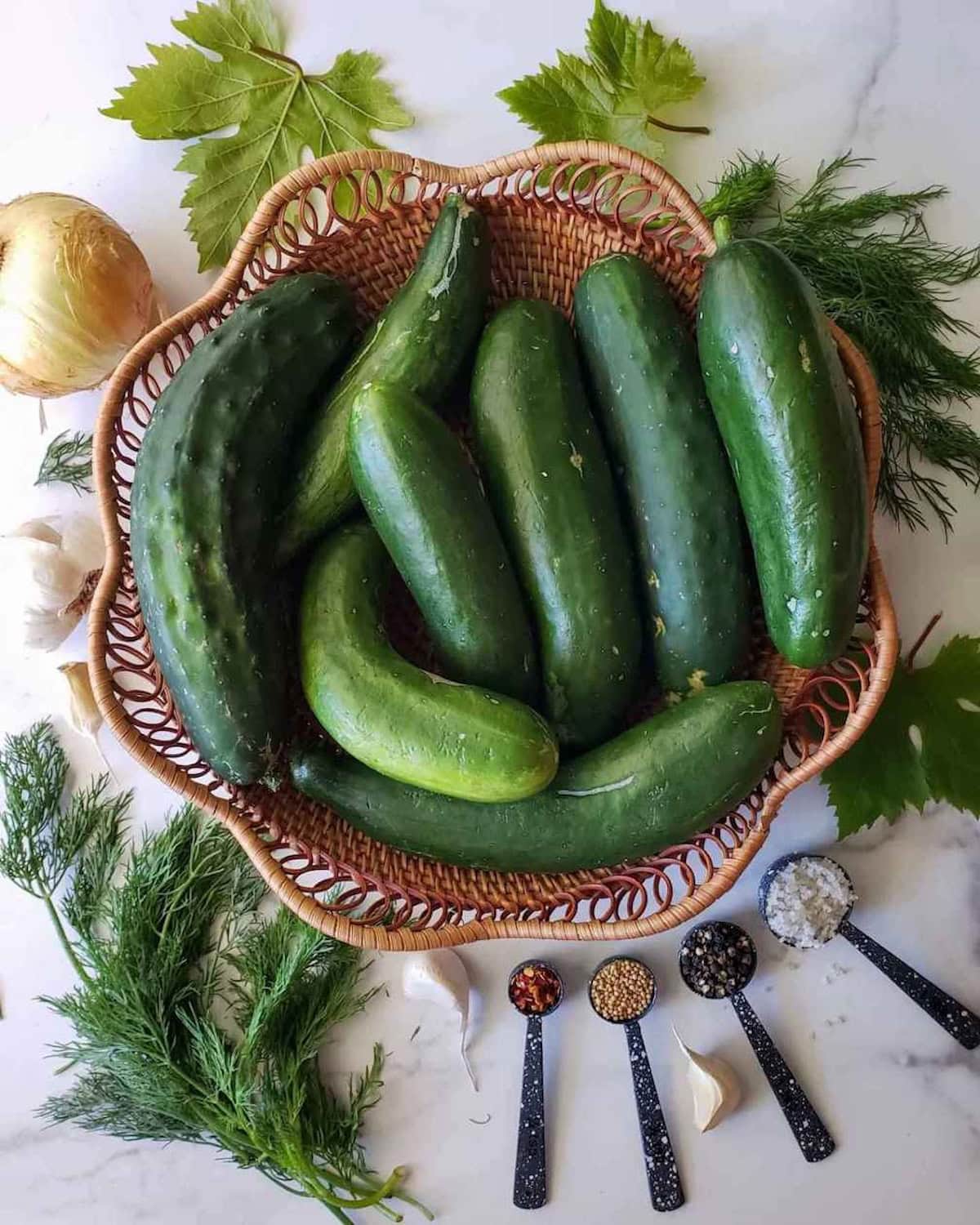 A wicker basket full of freshly harvested cucumbers, surrounding the basket are sprigs of fresh dill, onions, garlic, fresh grape leaf and spoonful measurements of chili flakes, mustard seeds, peppercorns, and sea salt.