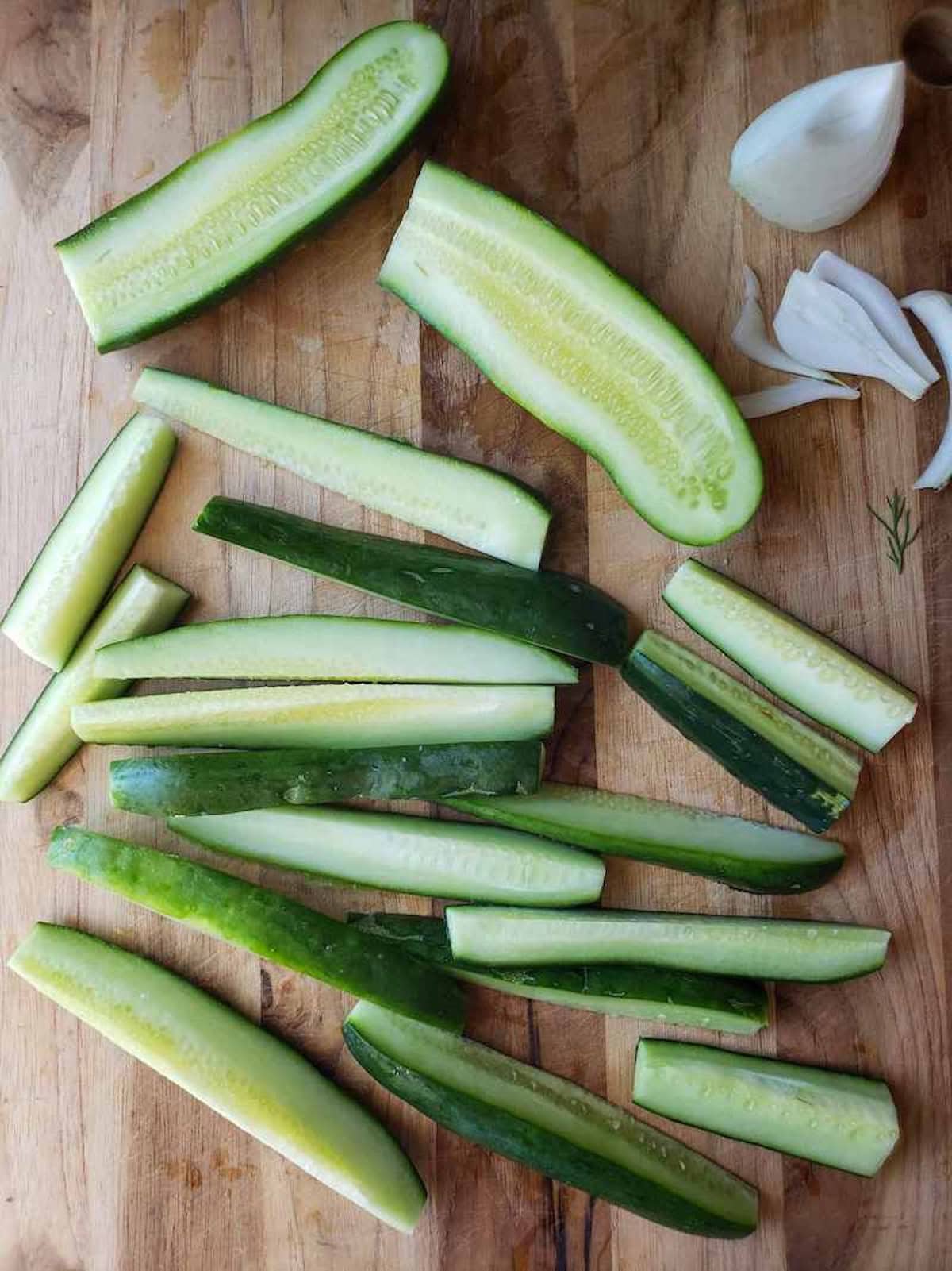 Looking down at a wooden cutting board with lengthwise cucumber slices, a cucumber sliced in half, and a few onion slices.