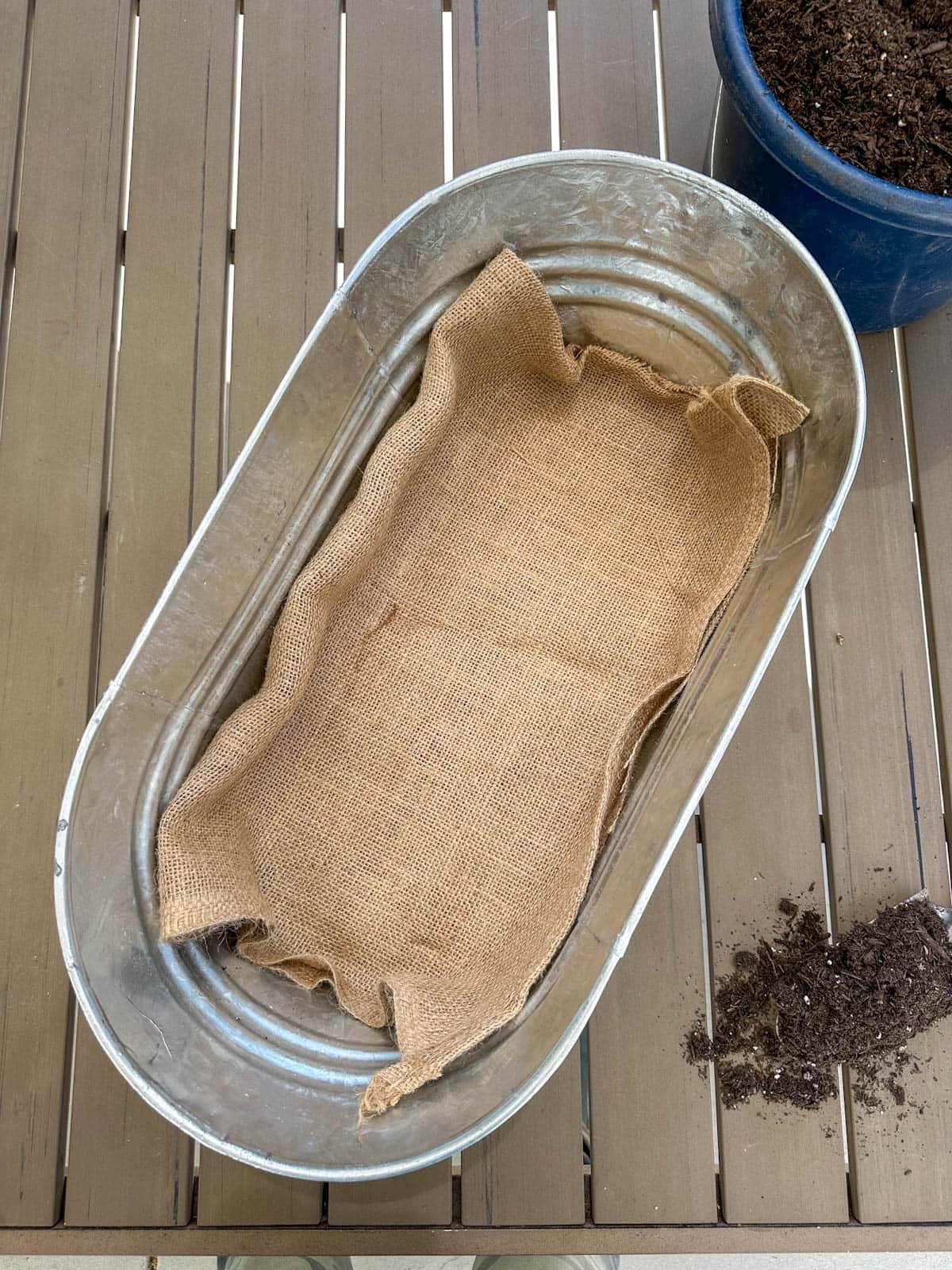 A long oval trough pot has a small section of burlap lining the bottom of it. A bucket of soil sits nearby.