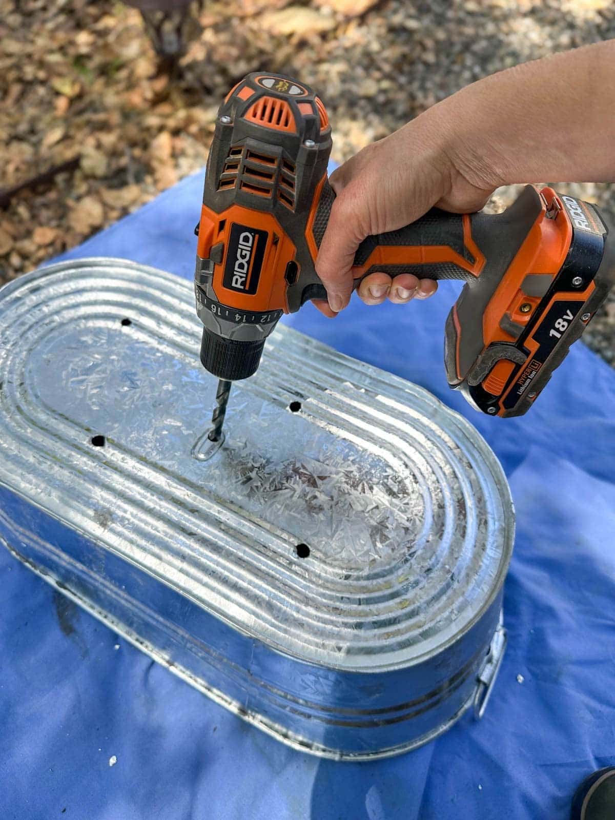 A metal trough is upside down while an electric drill is used to drill holes into the bottom of the container.