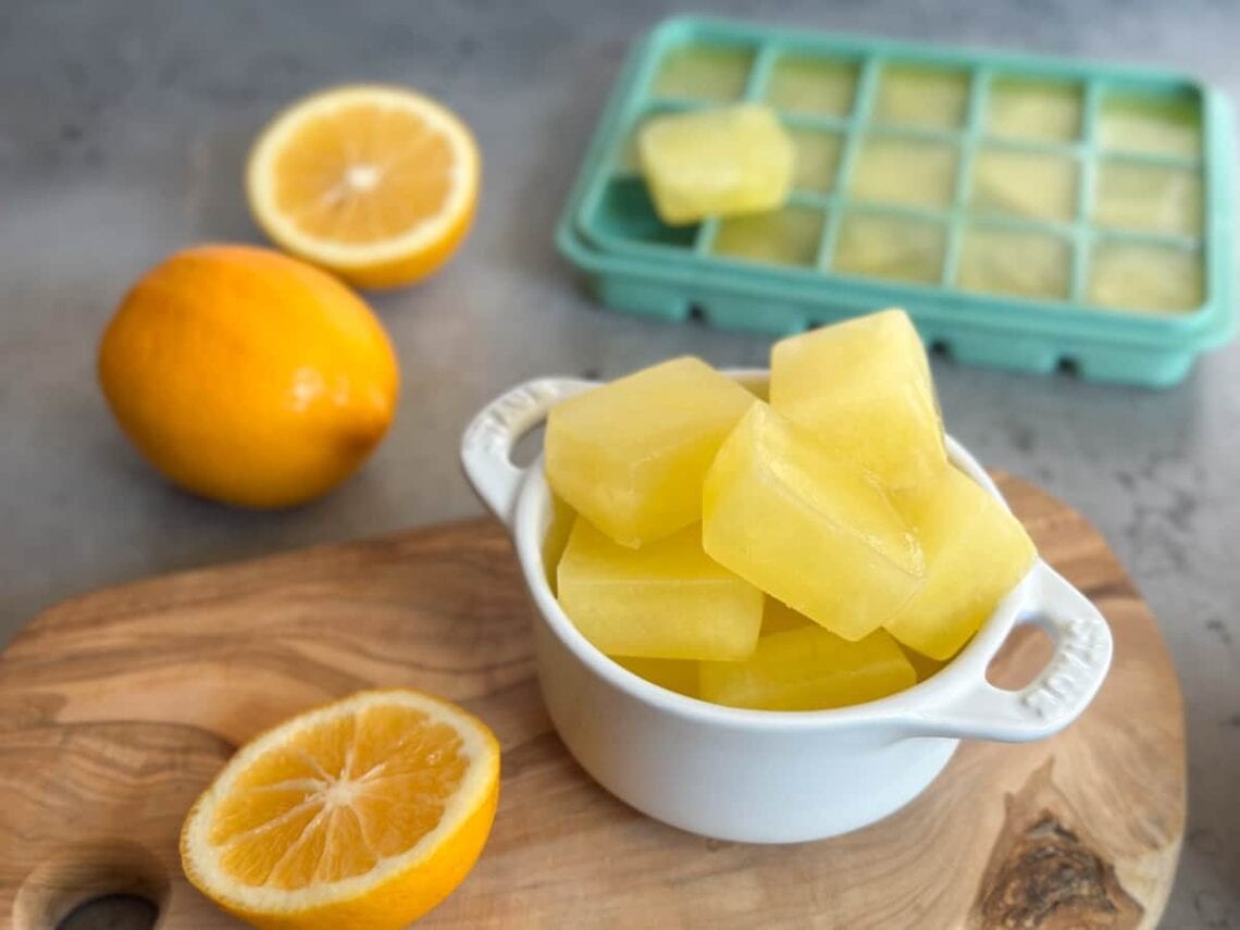 A small white ramekin filled with frozen lemon juice cubes. Surrounding the ramekin are one whole lemon along with one that has be cut in half as well as a silicone ice cube tray full of frozen lemon juice.