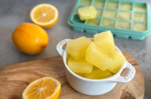 A small white ramekin filled with frozen lemon juice cubes. Surrounding the ramekin are one whole lemon along with one that has be cut in half as well as a silicone ice cube tray full of frozen lemon juice.