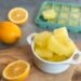 A small white ramekin filled with frozen lemon juice cubes. Surrounding the ramekin are one whole lemon along with one that has be cut in half as well as a silicone ice cube tray full of frozen lemon juice.