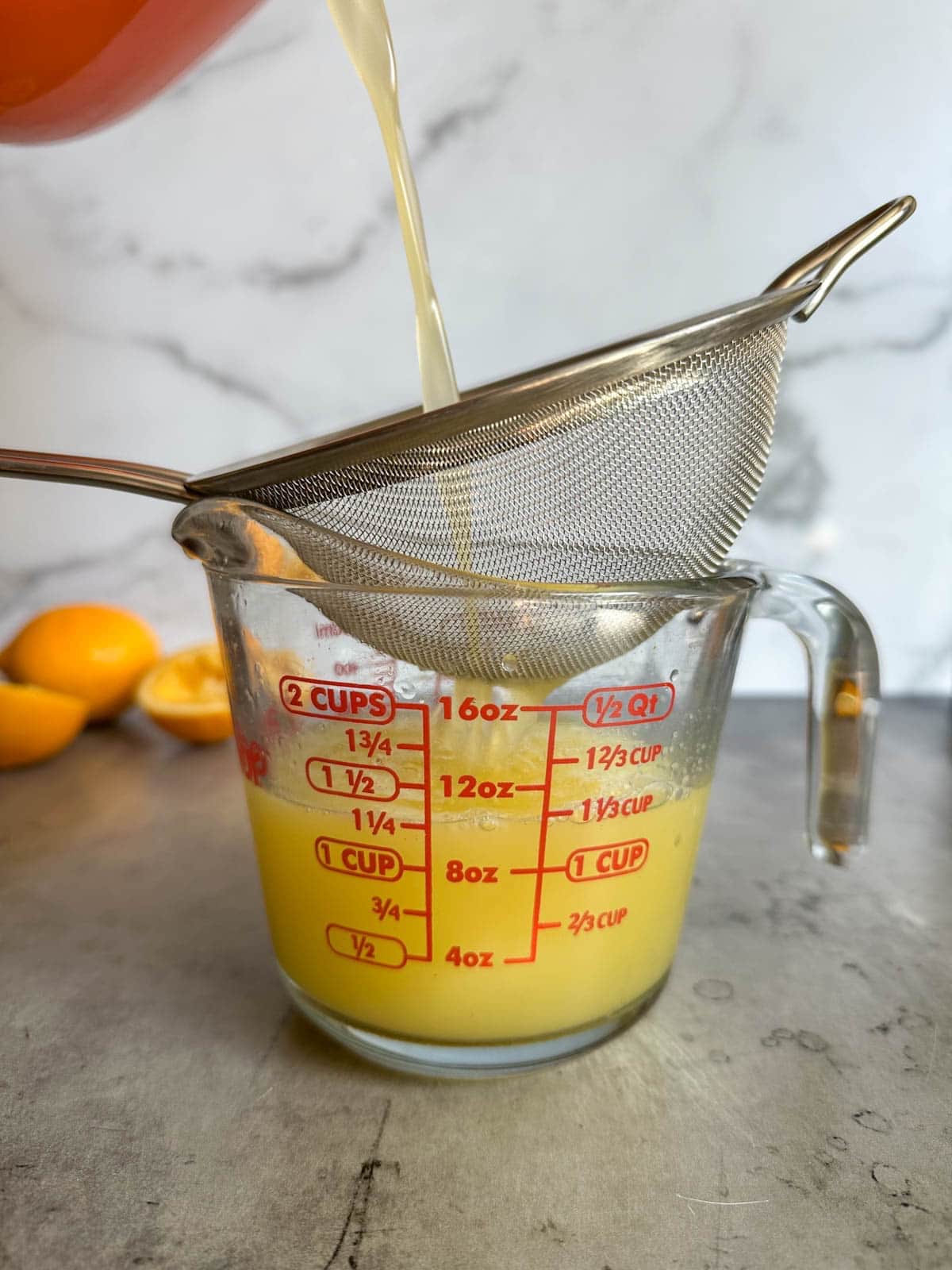 A fine mesh strainer is positioned above a liquid glass measuring cup as liquid is being poured from the top, through the strainer, into the measuring cup.