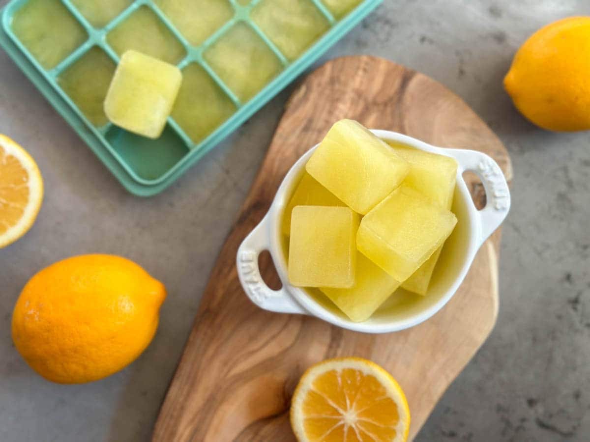 A small white ramekin is filled with frozen lemon juice cubes, sitting on a wooden cutting board. A few whole lemons and a lemon sliced in half are around the ramekin. A silicone ice cube tray full of frozen lemon juice is nearby.