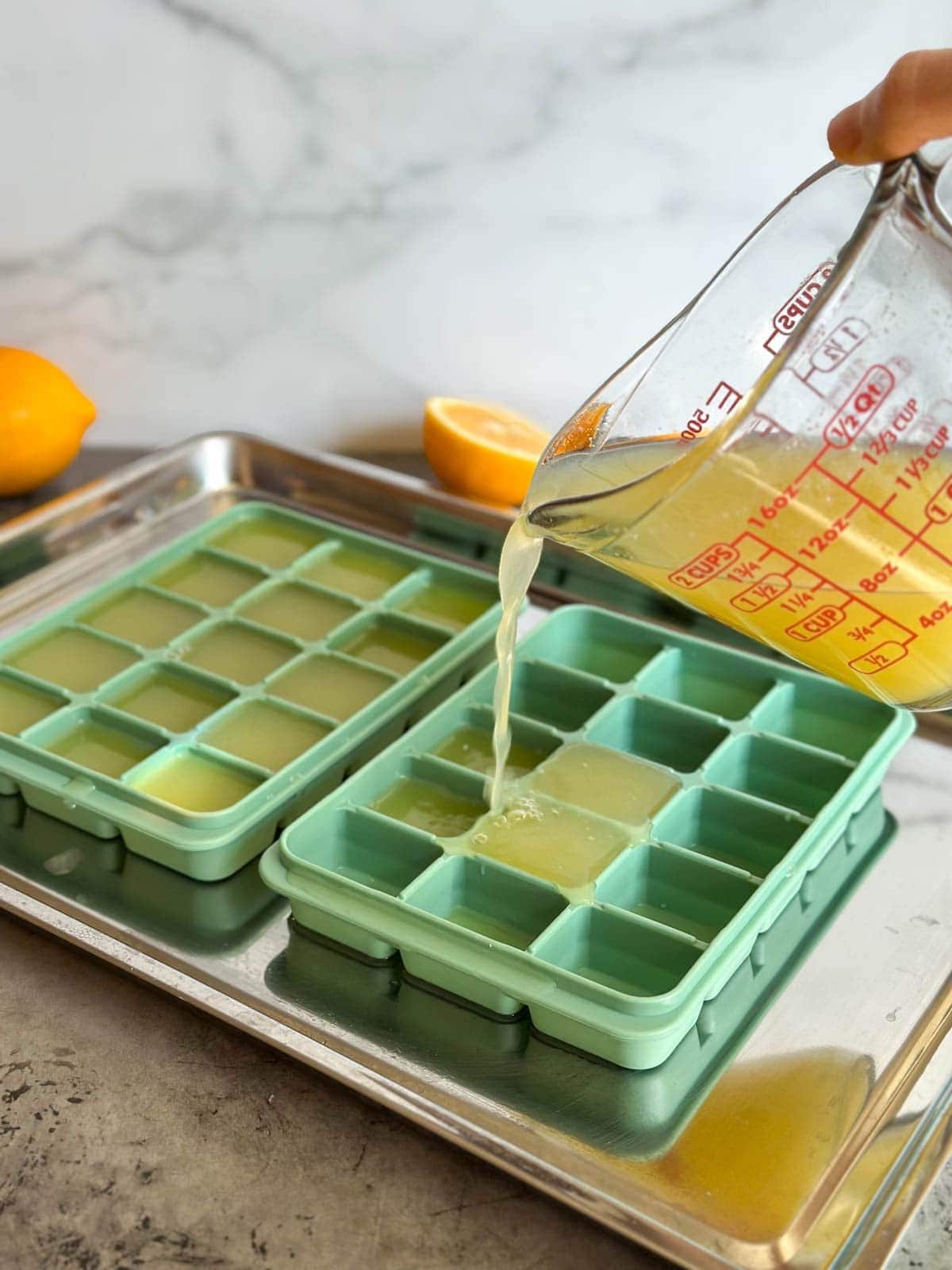 A liquid glass measuring cup is held above silicone trays, pouring liquid into each cell of the tray.